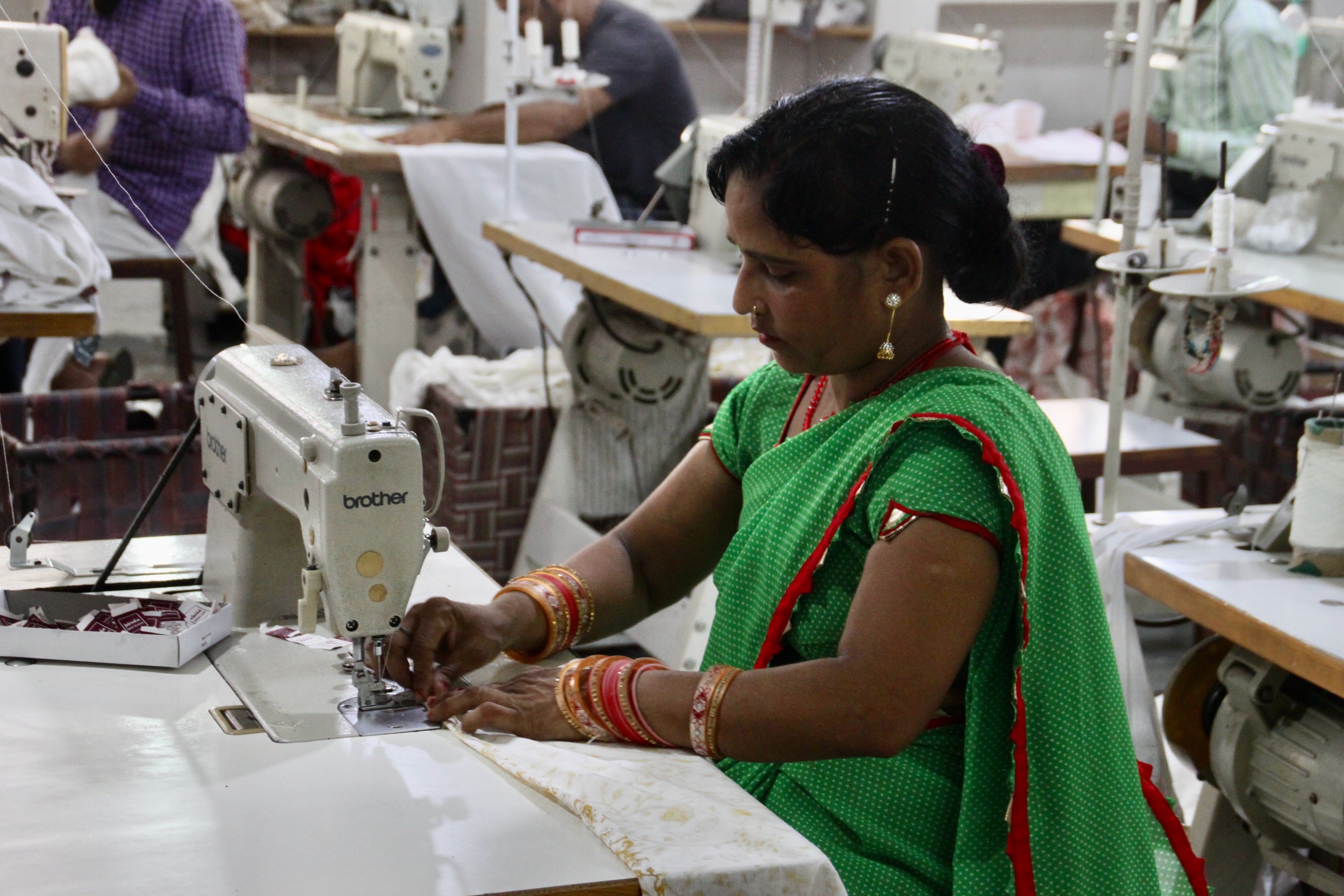 Vrouw met groene sari en goud/rode sieraden zit achter een witte naaimachine in de fabriek in India