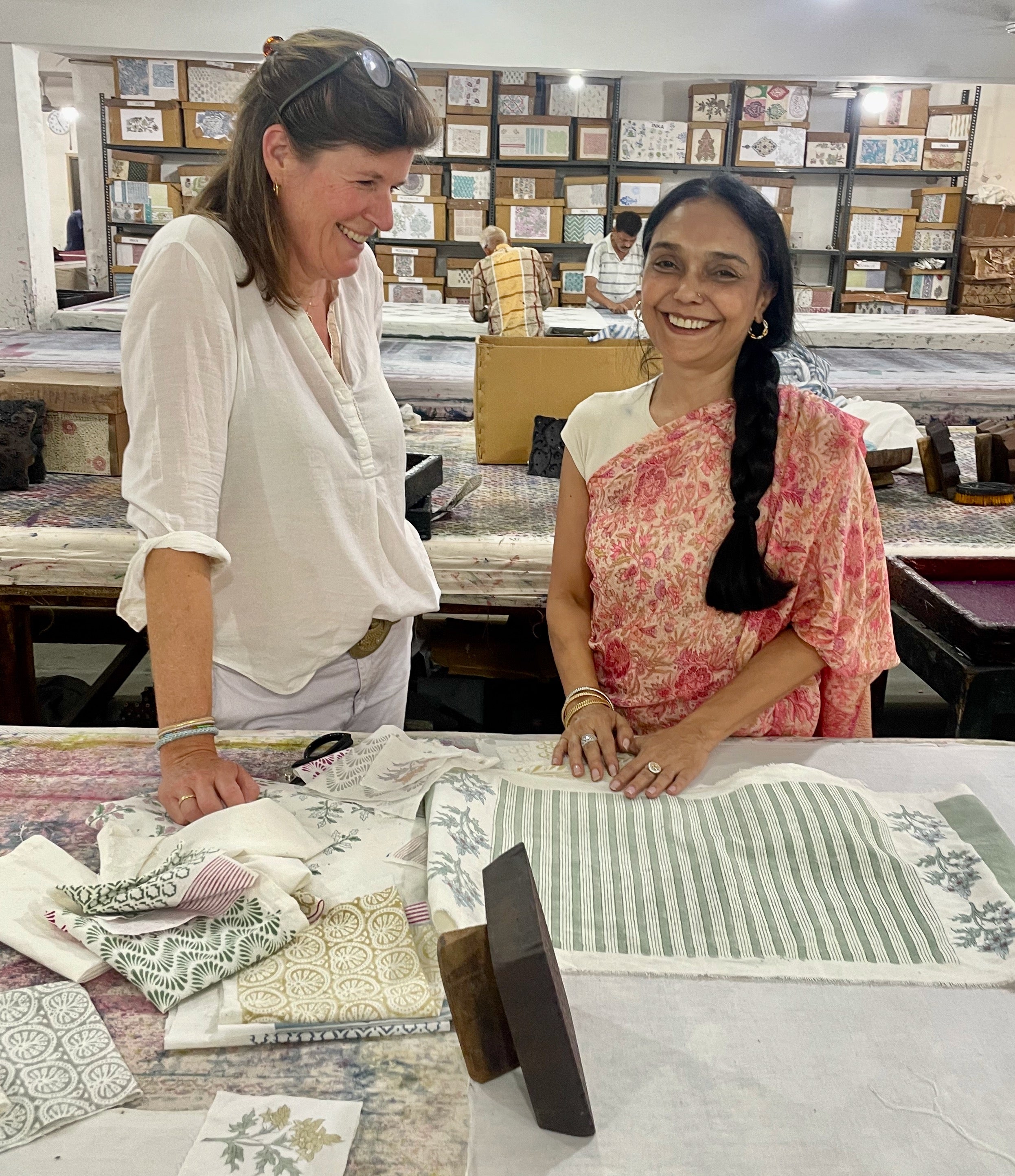Oprichter Karlijn staat naast ontwerper Indu, twee vrouwen die lachen, achter een tafel in hun fabriek met verschillende katoenen stoffen met gekleurde blockprints. 