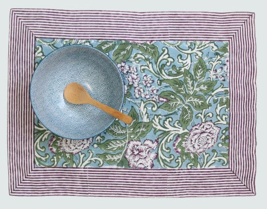 rechthoekige placemat van katoen met een rand van aubergine kleurige strepen. Binnen de rand een bloemenpatroon met aubergine kleurige bloemen, groene bladeren en groen en witte peasly-print. Op een blauwe achtergrond. Op de placemat staat een blauwe kom met houten lepel er in. 