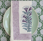 Servet met aubergine kleurige strepen en bloemenprint van bovenaf gezien, op een groen bordje met op de achtergrond een groen tafelkleed met bloemenprint. 